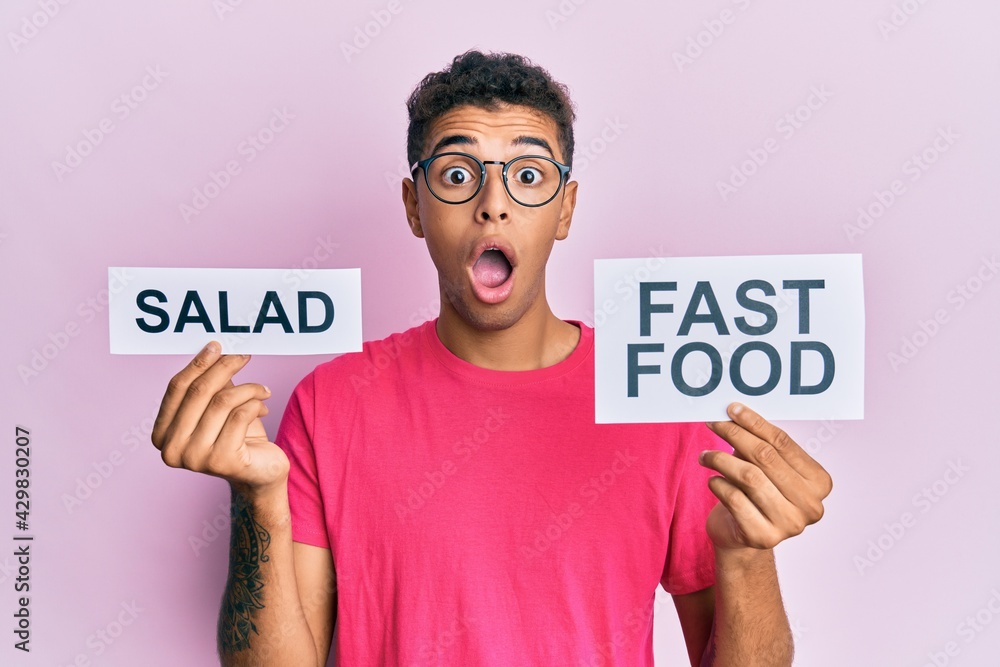 Sticker Young handsome african american man holding salad and fast food message paper afraid and shocked with surprise and amazed expression, fear and excited face.