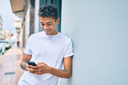 Young latin man smiling happy using smartphone leaning on the wall at the city.