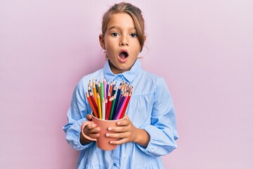 Little beautiful girl holding colored pencils afraid and shocked with surprise and amazed...