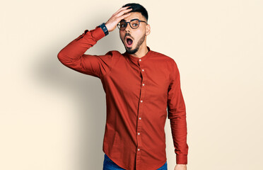 Young hispanic man with beard wearing business shirt and glasses surprised with hand on head for...