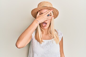 Young blonde girl wearing summer hat peeking in shock covering face and eyes with hand, looking...
