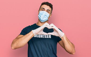 Hispanic young man wearing medical mask and volunteer t shirt smiling in love showing heart symbol...
