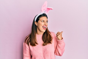 Obraz na płótnie Canvas Beautiful hispanic woman wearing cute easter bunny ears pointing thumb up to the side smiling happy with open mouth