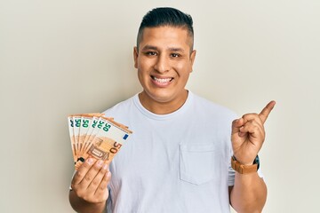 Young latin man holding bunch of 50 euro banknotes smiling happy pointing with hand and finger to the side