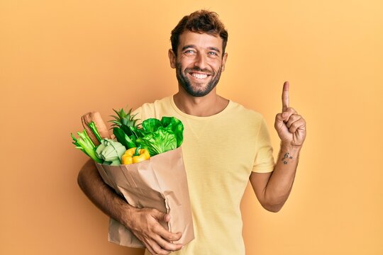 Handsome man with beard holding paper bag with bread and groceries smiling with an idea or question pointing finger with happy face, number one