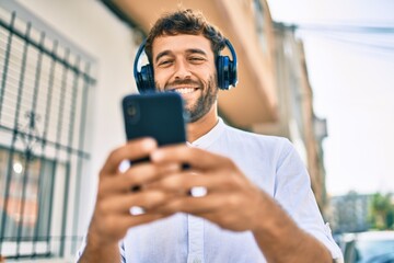 Handsome man with beard wearing casual white shirt on a sunny day smiling happy outdoors using smartphone listening to music wearing headphones