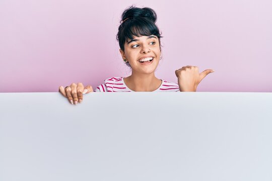 Young hispanic girl holding blank empty banner pointing thumb up to the side smiling happy with open mouth