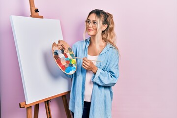 Beautiful young blonde woman standing drawing with palette by painter easel stand smiling looking to the side and staring away thinking.