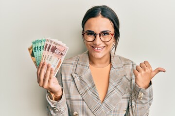 Young brunette woman holding 5000 south korean won banknotes pointing thumb up to the side smiling happy with open mouth