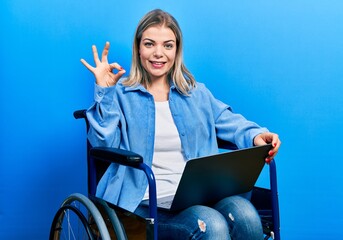 Beautiful caucasian woman sitting on wheelchair using laptop doing ok sign with fingers, smiling...