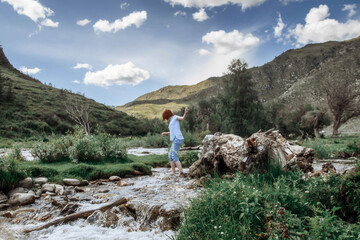 Girl crossing river against a backdrop of mountains and a blue sky with clouds. Young woman with red hair. Altay Republic, Siberia