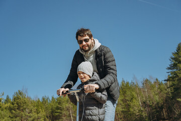 young bearded handsome man wearing hoodie, puffer jacket and sunglasses riding kick scooter with...