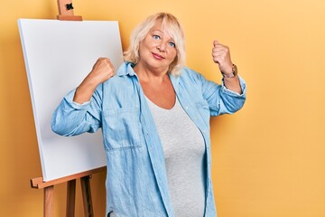 Middle age blonde woman standing by painter easel stand showing arms muscles smiling proud. fitness...