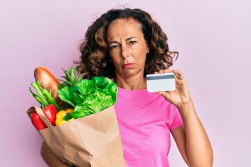 Middle age hispanic woman holding groceries and credit card clueless and confused expression. doubt concept.