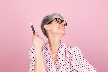 Happy positive excited stylish woman in dress and glasses on pink background holding heated tobacco...