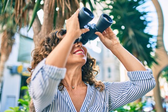 Middle age hispanic woman smiling happy looking for new opportunity using binoculars at the city.