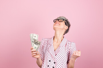 Stylish woman in dress and glasses on pink background holding money fan of 100 dollars  excited shocked do  winner gesture clenching fist