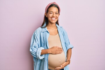 Beautiful hispanic woman expecting a baby, touching pregnant belly smiling and laughing hard out loud because funny crazy joke. - obrazy, fototapety, plakaty