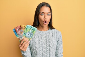 Beautiful hispanic woman holding australian dollars scared and amazed with open mouth for surprise, disbelief face