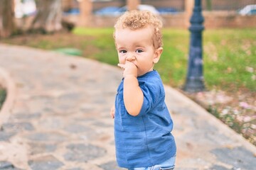 Sad little boy putting fingers on mouth touching gums because toothache at the park on a sunny day. Beautiful blonde hair male toddler in pain for new baby teeth outdoors