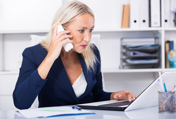 Smiling mature woman talking on mobile phone and using laptop in office