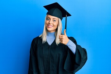 Beautiful blonde woman wearing graduation cap and ceremony robe doing happy thumbs up gesture with hand. approving expression looking at the camera showing success.