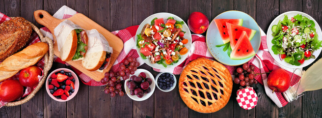 Summer picnic food table scene overhead view on a wood banner background. Variety of cold salads, sandwiches, fruit and treats.