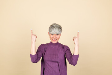 Stylish woman in purple casual on beige background happy excited cheerful point up with index finger