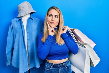 Young caucasian woman holding shopping bags serious face thinking about question with hand on chin,...