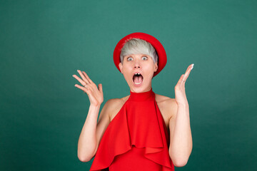 Studio portrait of stylish woman in red dress on green background happy look to camera shocked amazed surprised open mouth