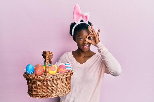 Young african american girl wearing cute easter bunny ears holding basket with painted eggs smiling happy doing ok sign with hand on eye looking through fingers
