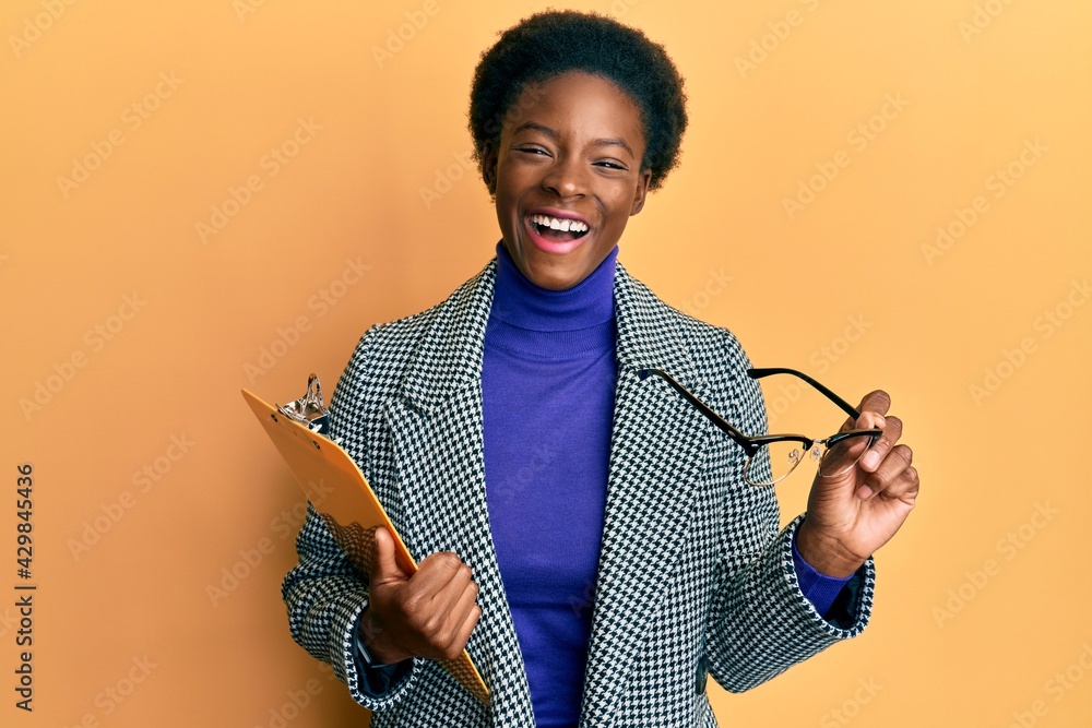 Poster Young african american girl holding clipboard and glasses smiling and laughing hard out loud because funny crazy joke.