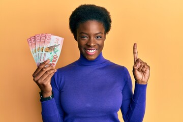 Young african american girl holding 100 new zealand dollars banknote smiling with an idea or question pointing finger with happy face, number one