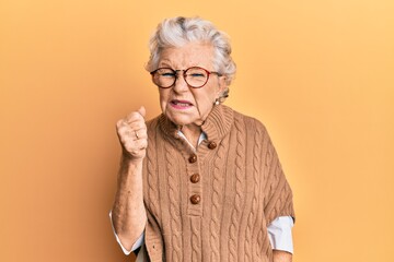 Senior grey-haired woman wearing casual clothes and glasses angry and mad raising fist frustrated...