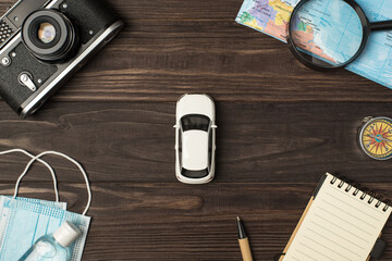 Top view photo of car model in the center camera map magnifier compass notebook pen medical masks and sanitizer on isolated wooden table background