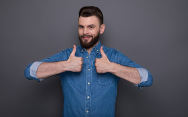 Modern happy excited young bearded man in jeans shirt shows thumbs up into the camera and having fun