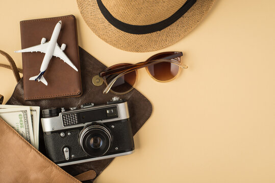 Top view photo of open leather bag with plane model on passport cover camera money sunglasses and sunhat on isolated beige background with copyspace