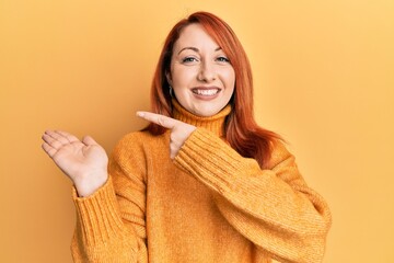 Beautiful redhead woman wearing casual winter sweater over yellow background amazed and smiling to the camera while presenting with hand and pointing with finger.