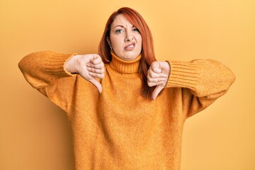 Beautiful redhead woman doing negative thumbs down gesture clueless and confused expression. doubt...