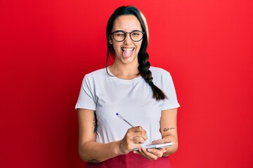 Young hispanic woman wearing waitress apron taking order sticking tongue out happy with funny...