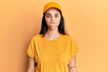 Young hispanic woman wearing delivery uniform and cap puffing cheeks with funny face. mouth...