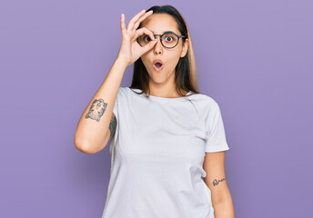 Young hispanic woman wearing casual white t shirt doing ok gesture shocked with surprised face, eye looking through fingers. unbelieving expression.