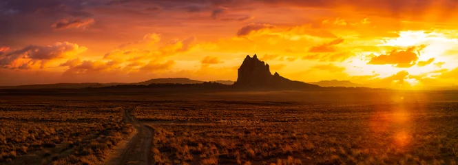 Foto op Plexiglas Opvallend panoramisch landschapsbeeld van een onverharde weg in de droge woestijn met een bergtop op de achtergrond. De kleurrijke Kunst van de Zonsonderganghemel geeft terug. Genomen op Shiprock, New Mexico, Verenigde Staten. © edb3_16