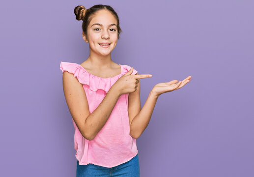 Beautiful brunette little girl wearing summer pink shirt amazed and smiling to the camera while presenting with hand and pointing with finger.