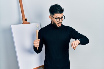 Handsome man with beard holding brushes close to easel stand looking at the watch time worried,...
