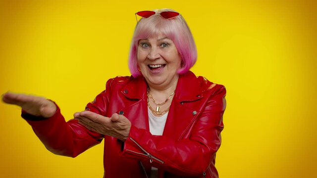 Mature stylish granny woman in red jacket showing gesture of wasting or throwing money around hand more tips dreaming about big profit body language. Senior grandmother isolated on yellow background