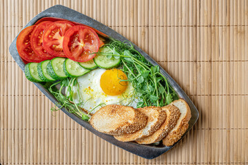 fried egg, toasted bread, cucumber and tomato slices and microgreens on a metal plate on bamboo straw surface, healthy breakfast concept, top view