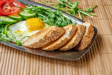 fried egg, toasted bread, cucumber and tomato slices and microgreens on a metal plate on bamboo straw surface, healthy breakfast concept