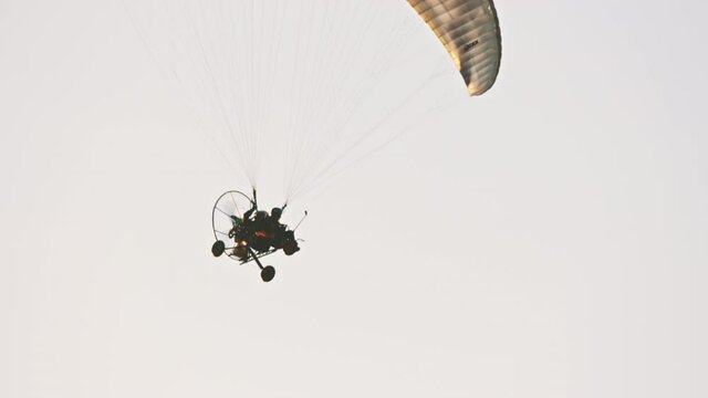 Flying Paramotor trike hanging under parachute. White sky background. Tandem motor powered paragliding at twilight. 