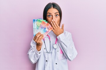 Beautiful hispanic woman wearing medical uniform holding swiss franc banknotes covering mouth with hand, shocked and afraid for mistake. surprised expression